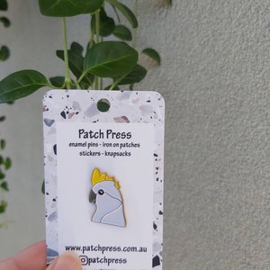 May include: White enamel pin of a cockatoo with a yellow crest. The pin is on a white card with the text "Patch Press" and "enamel pins - iron on patches stickers - knapsacks" and the website address "www.patchpress.com.au".