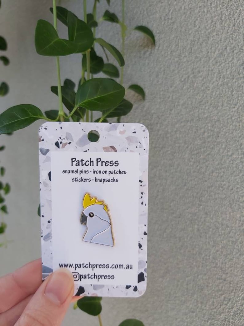May include: White enamel pin of a cockatoo with a yellow crest. The pin is on a white card with the text "Patch Press" and "enamel pins - iron on patches stickers - knapsacks" and the website address "www.patchpress.com.au".