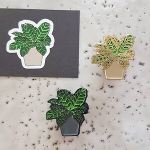 May include: Three plant-themed enamel pins and a sticker. The pins feature a green and gold Calathea plant design in a pot. One pin is gold, one is green and black, and the sticker is white with a black background.
