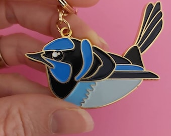 Peut inclure: Un porte-clés en métal doré représentant un oiseau bleu et noir avec un ventre blanc. L'oiseau a une tête bleue et un bec noir.