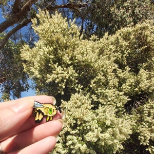 K&ouml;nnte beinhalten: Nahaufnahme einer goldenen Bienen-Brosche mit gr&uuml;nen Augen, die in einer Hand vor dem Hintergrund eines Busches mit wei&szlig;en Bl&uuml;ten gehalten wird.