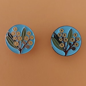 May include: Two round enamel pins with a blue background and a yellow flower design. The flowers are stylised and have a dark brown stem and leaves.