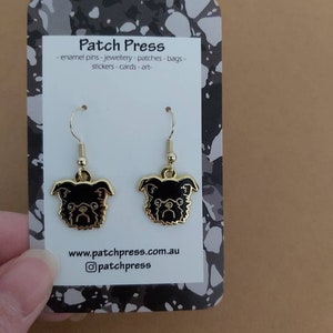 Puede incluir: Un par de pendientes de tono dorado con caras de perro de esmalte negro. Los pendientes est&aacute;n en una tarjeta blanca con el texto "Patch Press" y "www.patchpress.com.au" y el nombre de usuario de Instagram "@patchpress".