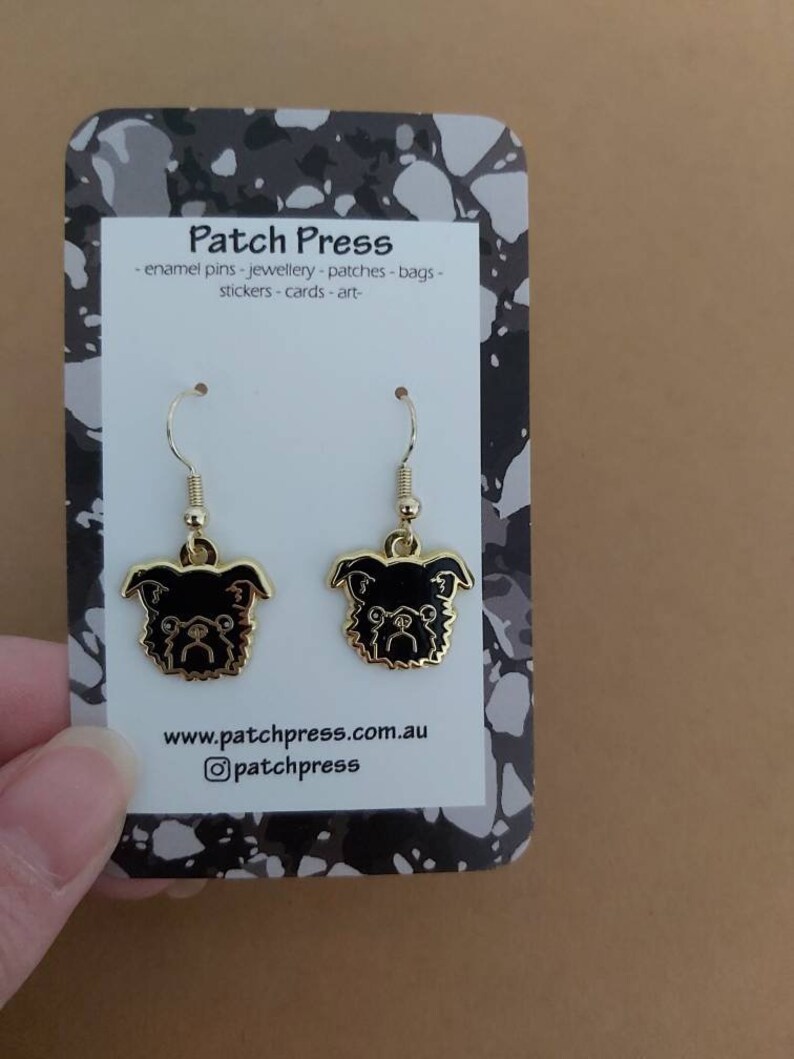 Puede incluir: Un par de pendientes de tono dorado con caras de perro de esmalte negro. Los pendientes est&aacute;n en una tarjeta blanca con el texto "Patch Press" y "www.patchpress.com.au" y el nombre de usuario de Instagram "@patchpress".