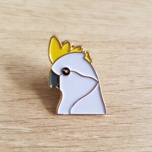 May include: A white and yellow enamel pin of a cockatoo's head. The pin has a gold finish.