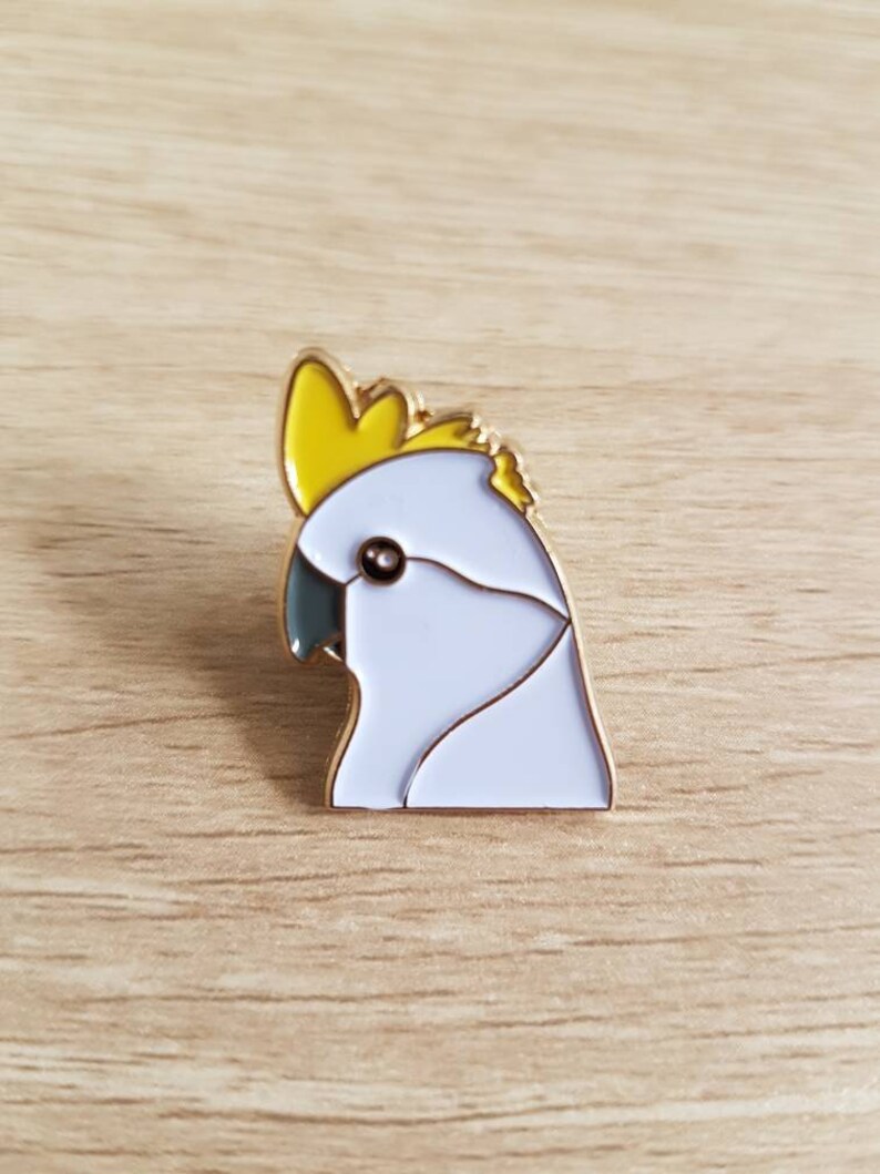 May include: A white and yellow enamel pin of a cockatoo's head. The pin has a gold finish.