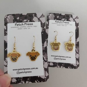 Puede incluir: Un par de pendientes de tono dorado con un dise&ntilde;o de pug marr&oacute;n. Los pendientes est&aacute;n en una tarjeta blanca con el texto "Patch Press" y una direcci&oacute;n de sitio web.