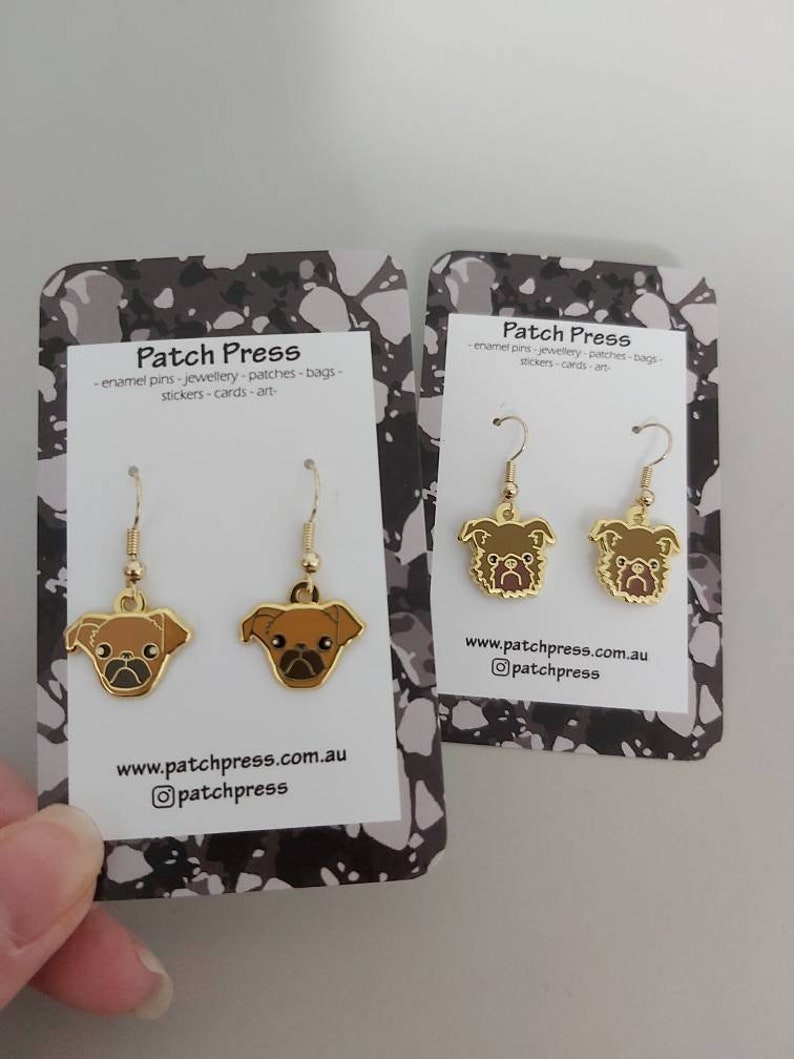 Puede incluir: Un par de pendientes de tono dorado con un dise&ntilde;o de pug marr&oacute;n. Los pendientes est&aacute;n en una tarjeta blanca con el texto "Patch Press" y una direcci&oacute;n de sitio web.