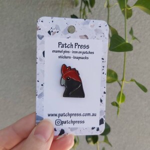 May include: A black and red enamel pin of a cockatoo bird. The pin is on a white card with the text "Patch Press" and "enamel pins - iron on patches stickers - knapsacks" and the website address "www.patchpress.com.au".