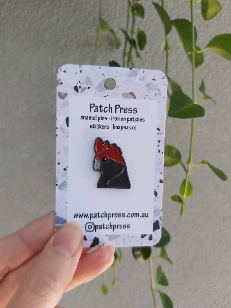 May include: A black and red enamel pin of a cockatoo bird. The pin is on a white card with the text "Patch Press" and "enamel pins - iron on patches stickers - knapsacks" and the website address "www.patchpress.com.au".