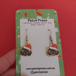 May include: A pair of gold-tone earrings featuring a cartoon Wombat wearing a Santa hat. The earrings are on a white card with the text "Patch Press - enamel pins - jewellery - patches - bags - stickers - cards - art - www.patchpress.com.au @patchpress"