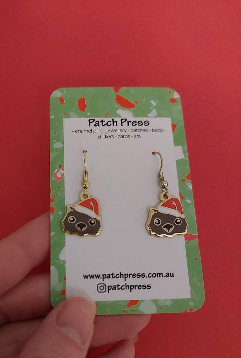 May include: A pair of gold-tone earrings featuring a cartoon Wombat wearing a Santa hat. The earrings are on a white card with the text "Patch Press - enamel pins - jewellery - patches - bags - stickers - cards - art - www.patchpress.com.au @patchpress"