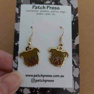 Puede incluir: Un par de pendientes de tono dorado con una ilustraci&oacute;n de dibujos animados de un perro grif&oacute;n de Bruselas marr&oacute;n y negro. Los pendientes est&aacute;n en una tarjeta blanca con el texto "Patch Press - enamel pins - jewellery - patches - bags - stickers - cards - art - www.patchpress.com.au @patchpress".