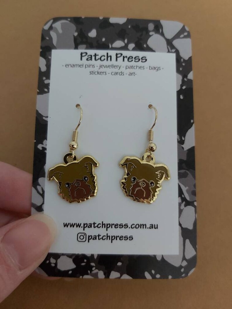 Puede incluir: Un par de pendientes de tono dorado con una ilustraci&oacute;n de dibujos animados de un perro grif&oacute;n de Bruselas marr&oacute;n y negro. Los pendientes est&aacute;n en una tarjeta blanca con el texto "Patch Press - enamel pins - jewellery - patches - bags - stickers - cards - art - www.patchpress.com.au @patchpress".