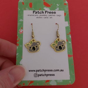 May include: A pair of gold dangle earrings featuring a koala bear with a green holly wreath. The earrings are displayed on a white card with the text "Patch Press - enamel pins - jewellery - patches - bags - stickers - cards - art - www.patchpress.com.au @patchpress"