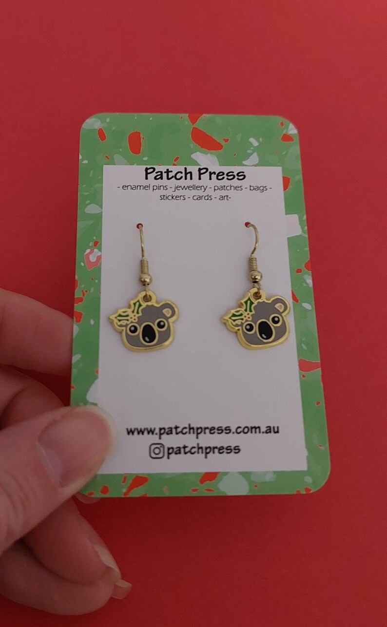 May include: A pair of gold dangle earrings featuring a koala bear with a green holly wreath. The earrings are displayed on a white card with the text "Patch Press - enamel pins - jewellery - patches - bags - stickers - cards - art - www.patchpress.com.au @patchpress"