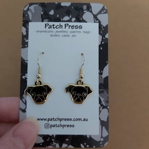 Puede incluir: Pendientes de aro dorados con caras de carlino de esmalte negro. Los pendientes est&aacute;n en una tarjeta blanca con el texto "Patch Press" y "www.patchpress.com.au" y "@patchpress".
