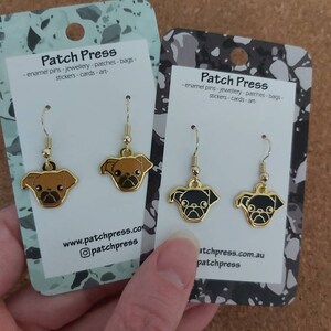 Puede incluir: Dos pares de pendientes de tono dorado con dijes de perro pug. Los dijes son de esmalte negro y marr&oacute;n. Los pendientes se muestran en tarjetas blancas con el texto "Patch Press - enamel pins - jewellery - patches - bags - stickers - cards - art - www.patchpress.com @patchpress" y "Patch Press - enamel pins - jewellery - patches - bags - stickers - cards - art - atchpress.com.au patchpress".