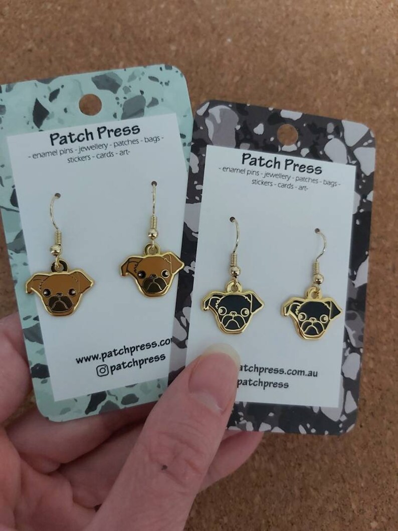 Puede incluir: Dos pares de pendientes de tono dorado con dijes de perro pug. Los dijes son de esmalte negro y marr&oacute;n. Los pendientes se muestran en tarjetas blancas con el texto "Patch Press - enamel pins - jewellery - patches - bags - stickers - cards - art - www.patchpress.com @patchpress" y "Patch Press - enamel pins - jewellery - patches - bags - stickers - cards - art - atchpress.com.au patchpress".