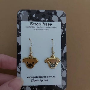 Puede incluir: Pendientes colgantes de tono dorado con un dise&ntilde;o de pug marr&oacute;n. Los pendientes se muestran en una tarjeta con el texto "Patch Press - enamel pins - jewellery - patches - bags - stickers - cards - art - www.patchpress.com.au @patchpress"