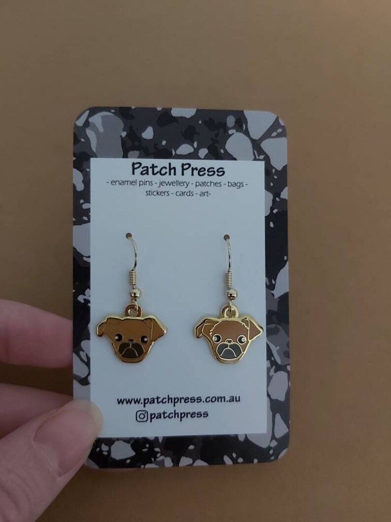 Puede incluir: Pendientes colgantes de tono dorado con un dise&ntilde;o de pug marr&oacute;n. Los pendientes se muestran en una tarjeta con el texto "Patch Press - enamel pins - jewellery - patches - bags - stickers - cards - art - www.patchpress.com.au @patchpress"