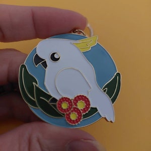 May include: A white cockatoo with a yellow crest is perched on a branch with red flowers. The cockatoo is surrounded by green leaves and a blue background. The image is a metal pin.