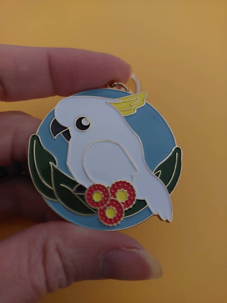 May include: A white cockatoo with a yellow crest is perched on a branch with red flowers. The cockatoo is surrounded by green leaves and a blue background. The image is a metal pin.