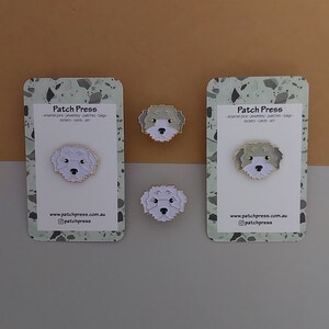 May include: Two white cards with a green and white speckled background. The cards have the text "Patch Press" and the text "enamel pins-jewellery-patches-bags-stickers-cards-art" on them. There are three small enamel pins of a white dog with black eyes and a black nose. The pins are on the cards and on a brown surface.