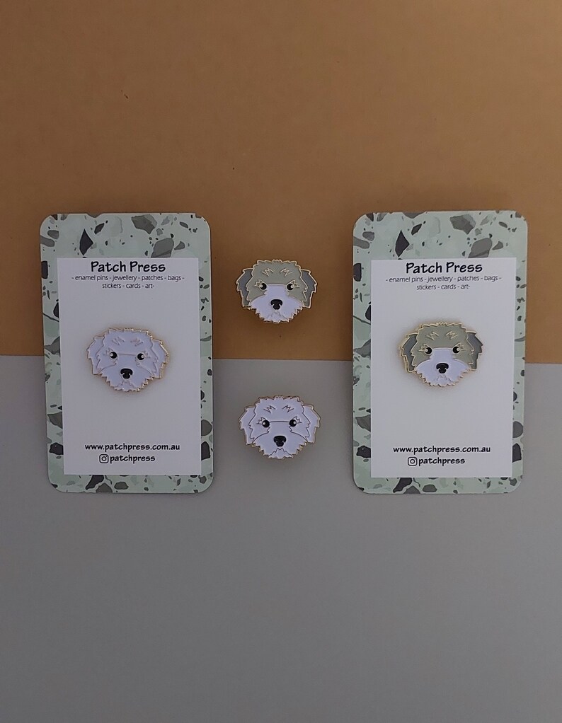 May include: Two white cards with a green and white speckled background. The cards have the text "Patch Press" and the text "enamel pins-jewellery-patches-bags-stickers-cards-art" on them. There are three small enamel pins of a white dog with black eyes and a black nose. The pins are on the cards and on a brown surface.