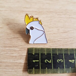 May include: A gold-colored enamel pin featuring a white cockatoo with a yellow crest. The pin is approximately 1 inch in size.