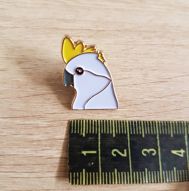 May include: A gold-colored enamel pin featuring a white cockatoo with a yellow crest. The pin is approximately 1 inch in size.