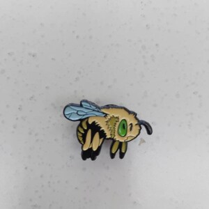 K&ouml;nnte beinhalten: Eine Emaillestifte mit einer Cartoon-Biene mit einem gr&uuml;nen Auge. Die Biene ist gelb und schwarz mit einem blau-wei&szlig; gestreiften Hinterleib und fliegt.