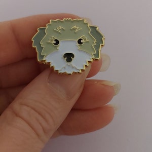 May include: A gold-colored enamel pin featuring a cartoon illustration of a dog's face. The dog has brown fur, a white muzzle, and black eyes.
