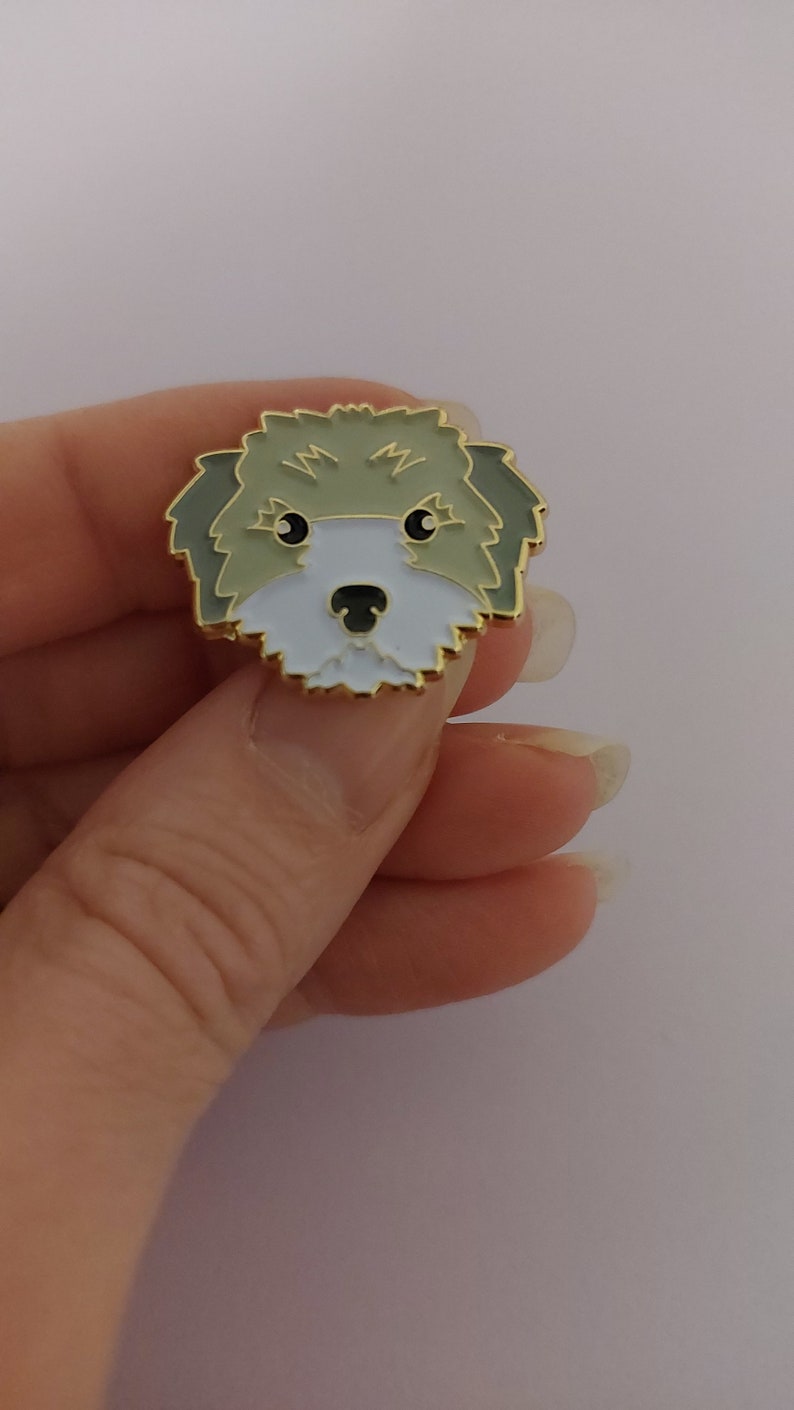 May include: A gold-colored enamel pin featuring a cartoon illustration of a dog's face. The dog has brown fur, a white muzzle, and black eyes.