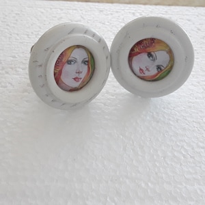 Op de afbeelding: Twee witte kastknoppen met een vintage ontwerp. Elke knop heeft een kleine, ronde afbeelding van een vrouw met rode lippen, blond haar en een bloemenkrans. De knoppen hebben een getextureerd, licht verweerd uiterlijk.
