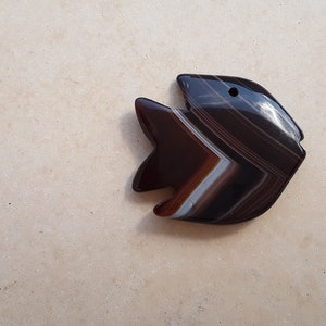 Puede incluir: Un colgante de pez de ágata negro y blanco con un agujero para un cordón o cadena. El pez está tallado en una piedra de ágata bandeada con un patrón natural.