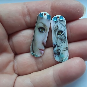 May include: Two oval-shaped pendants with a white background and a glittery finish. One pendant features a close-up of a woman's eye and lips, while the other pendant features a close-up of a snow leopard's eye. Both pendants have blue rhinestones at the top.