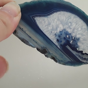 May include: A blue and white agate slice with a natural, irregular shape. The agate has a white center with blue and green bands radiating outward.