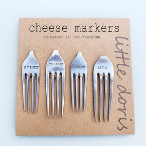 May include: Four stainless steel cheese markers on a brown card. Each fork is labelled with a cheese descriptor: "Stinky", "Creamy", "Tangy", and "Mild". The card reads "cheese markers created in twickenham" and "little doris".
