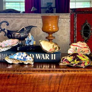 Pode incluir: Uma coleção de quatro conchas de ostra decorativas com bordas pintadas a ouro, cada uma apresentando padrões de decoupage únicos, incluindo motivos florais, listrados e azuis e brancos, dispostas sobre um livro intitulado World War II.