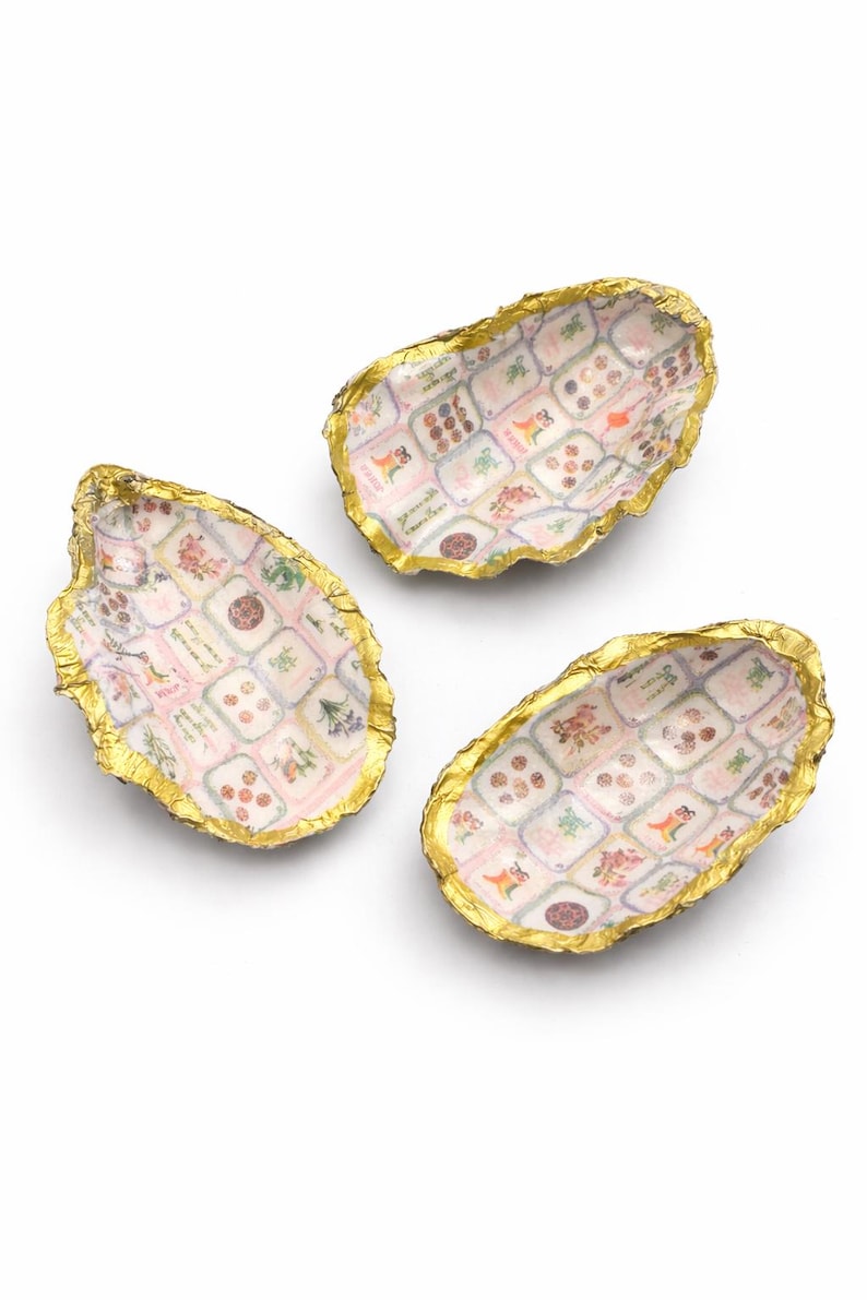 May include: Three oyster shell dishes with gold-painted edges. The interior of each shell features a grid pattern of small, colorful botanical and floral illustrations in shades of pink, green, and brown against a white background.