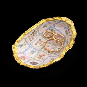 May include: An oval-shaped trinket dish with a textured, metallic gold rim and a white interior featuring a grid of small, colorful floral and botanical illustrations. Three gold-toned rings rest inside the dish, including one textured band.