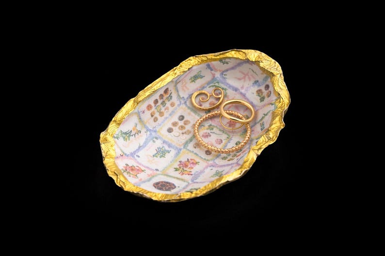 May include: An oval-shaped trinket dish with a textured, metallic gold rim and a white interior featuring a grid of small, colorful floral and botanical illustrations. Three gold-toned rings rest inside the dish, including one textured band.