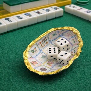May include: A small, oval-shaped bowl with a crinkled gold-colored rim and an interior featuring a grid of various floral and patterned illustrations. Three white six-sided dice with black pips are resting inside the bowl on a green surface.