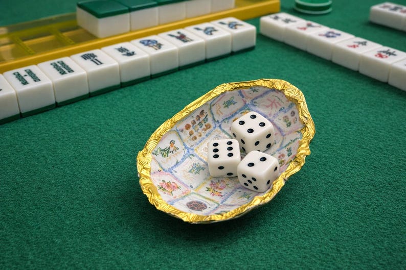 May include: A small, oval-shaped bowl with a crinkled gold-colored rim and an interior featuring a grid of various floral and patterned illustrations. Three white six-sided dice with black pips are resting inside the bowl on a green surface.