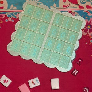 May include: A close-up shot of a board game set on a red surface. The set includes a light green game board with a scalloped edge and a grid of squares with blue numbers and letters. Scattered around are game tiles and red dice.