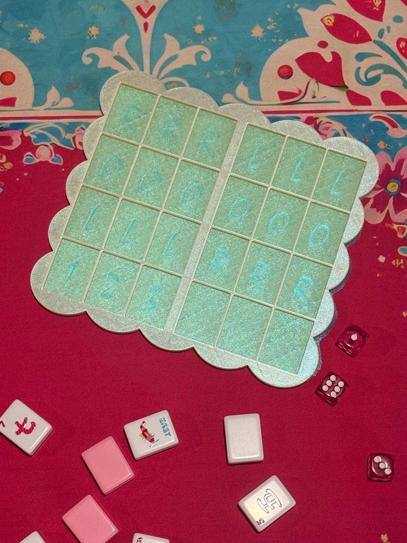 May include: A close-up shot of a board game set on a red surface. The set includes a light green game board with a scalloped edge and a grid of squares with blue numbers and letters. Scattered around are game tiles and red dice.