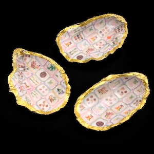 May include: Three oyster shells with gold-leafed edges and a decoupage interior featuring a grid of small, colorful floral and botanical illustrations on a light pink and white background, arranged in a cluster against a black backdrop.