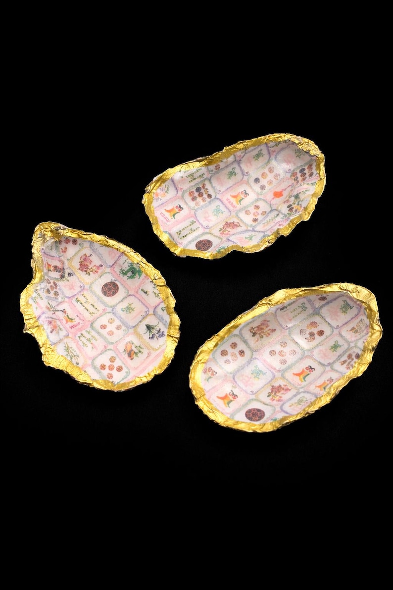 May include: Three oyster shells with gold-leafed edges and a decoupage interior featuring a grid of small, colorful floral and botanical illustrations on a light pink and white background, arranged in a cluster against a black backdrop.