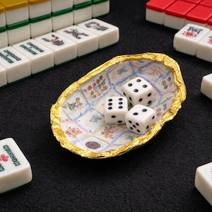 May include: An oval-shaped decorative dish with a gold-toned, textured rim and a white interior featuring a grid of small, colorful floral and patterned illustrations. Three white six-sided dice with black pips rest inside the dish.